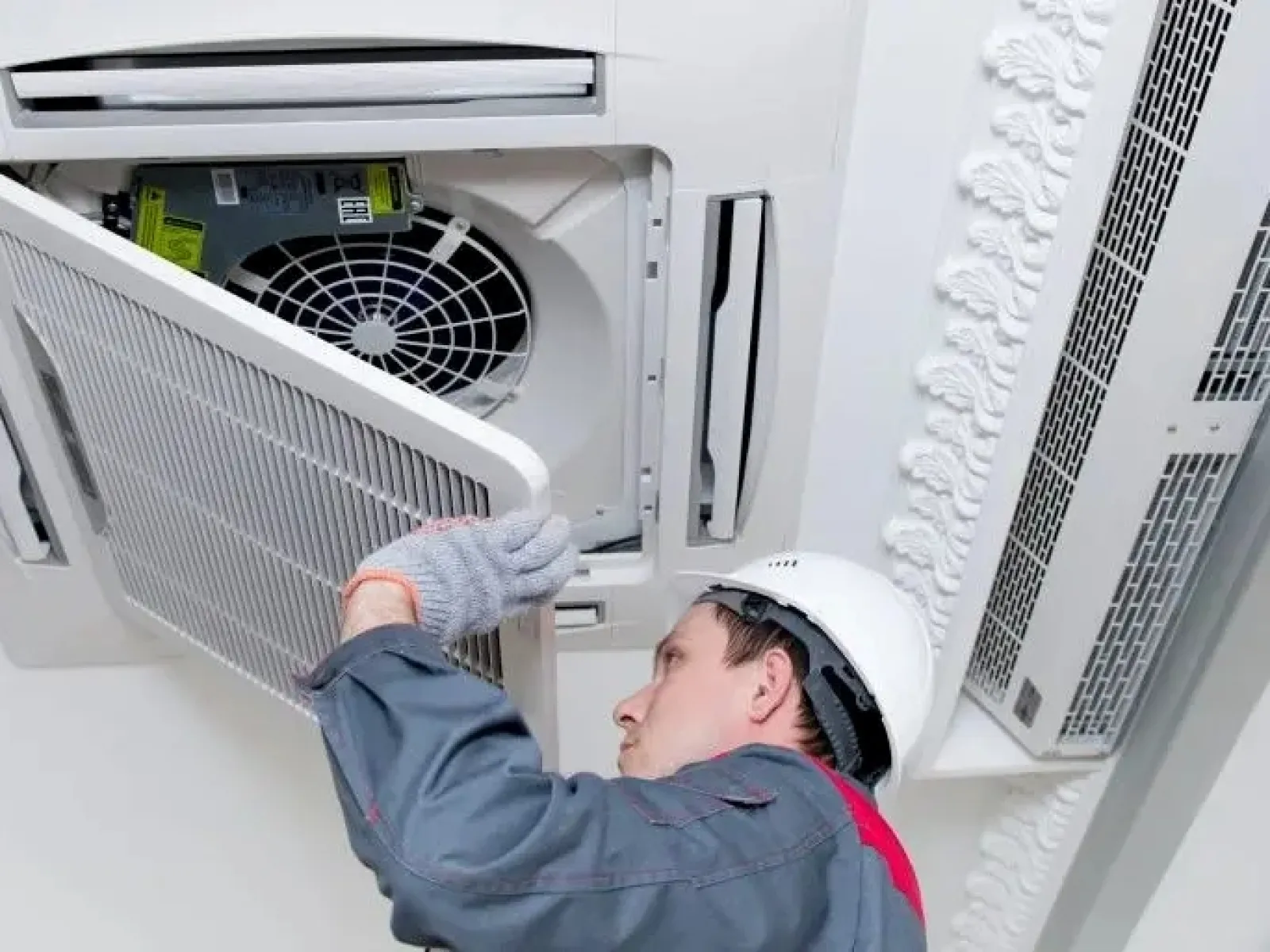 Technician in safety helmet and gloves installing or cleaning ceiling air conditioner filter in office space.