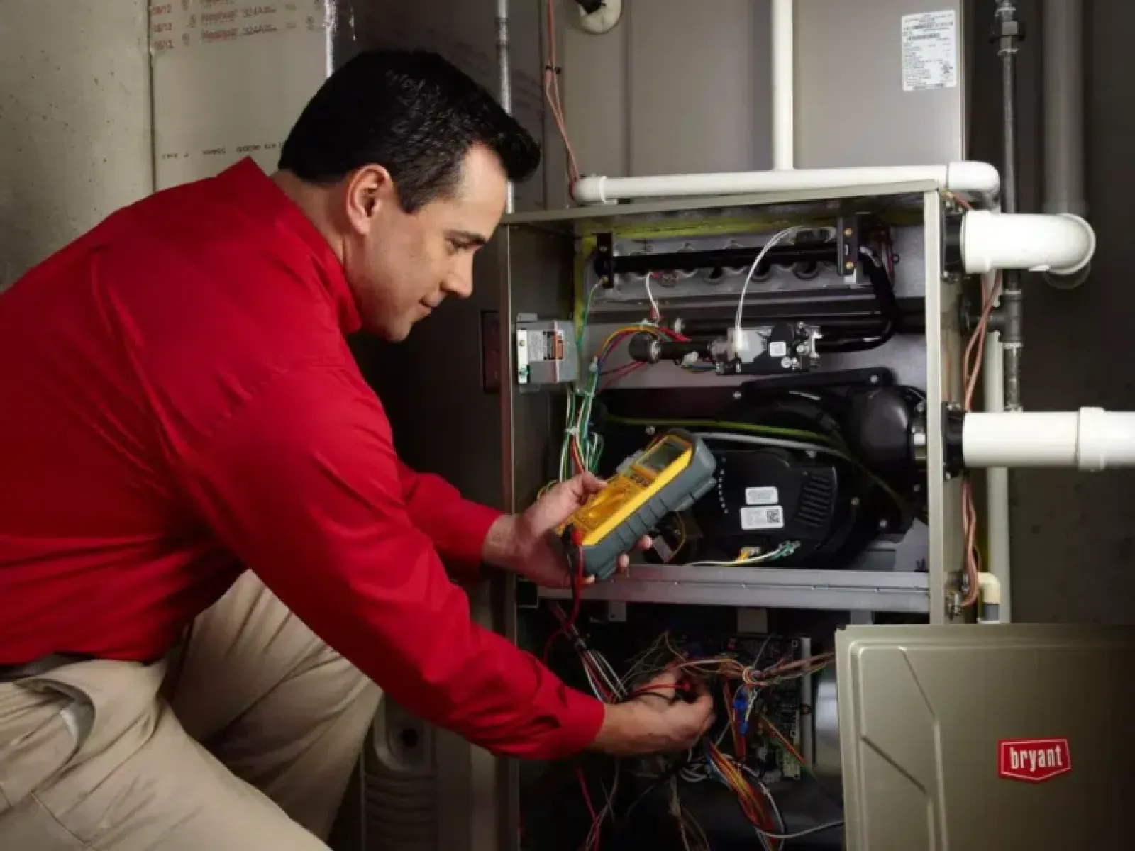 Technician in red shirt servicing HVAC unit using a multimeter to check electrical connections indoors.