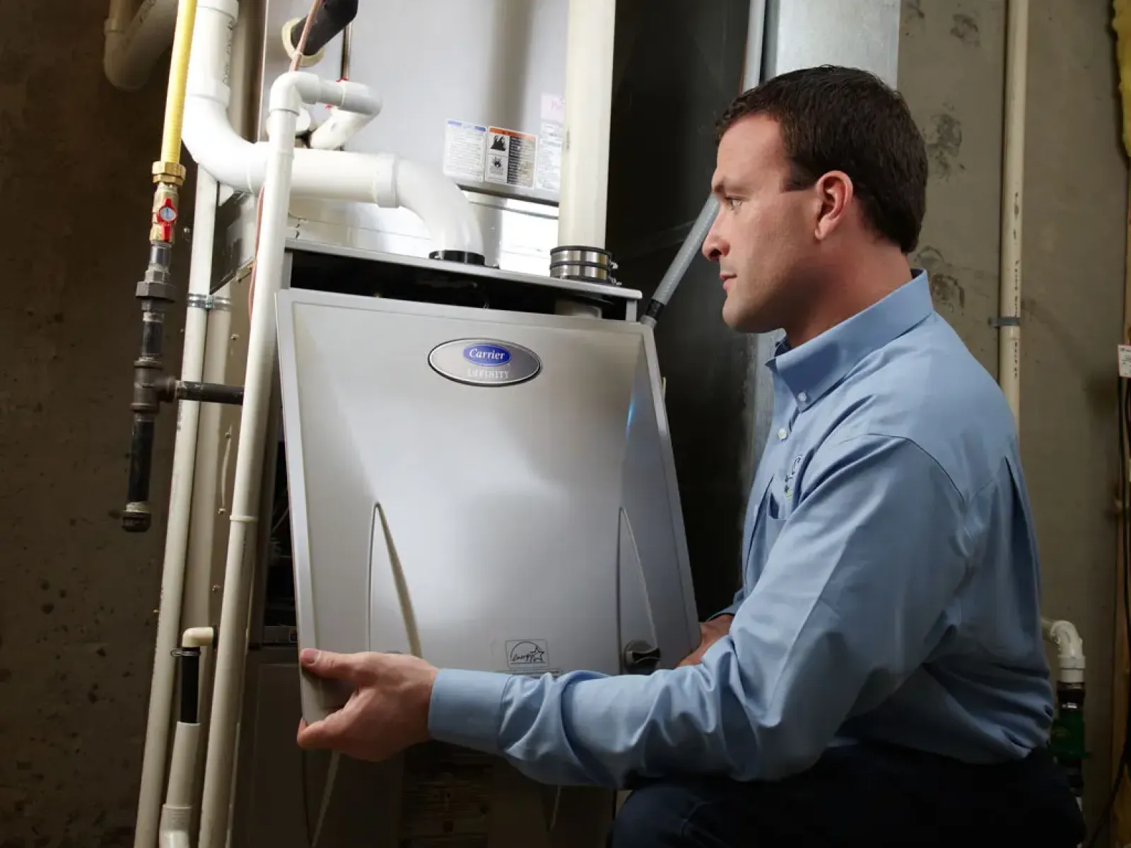 Technician inspecting a Carrier Infinity HVAC unit in a basement with connected pipes and ducts.