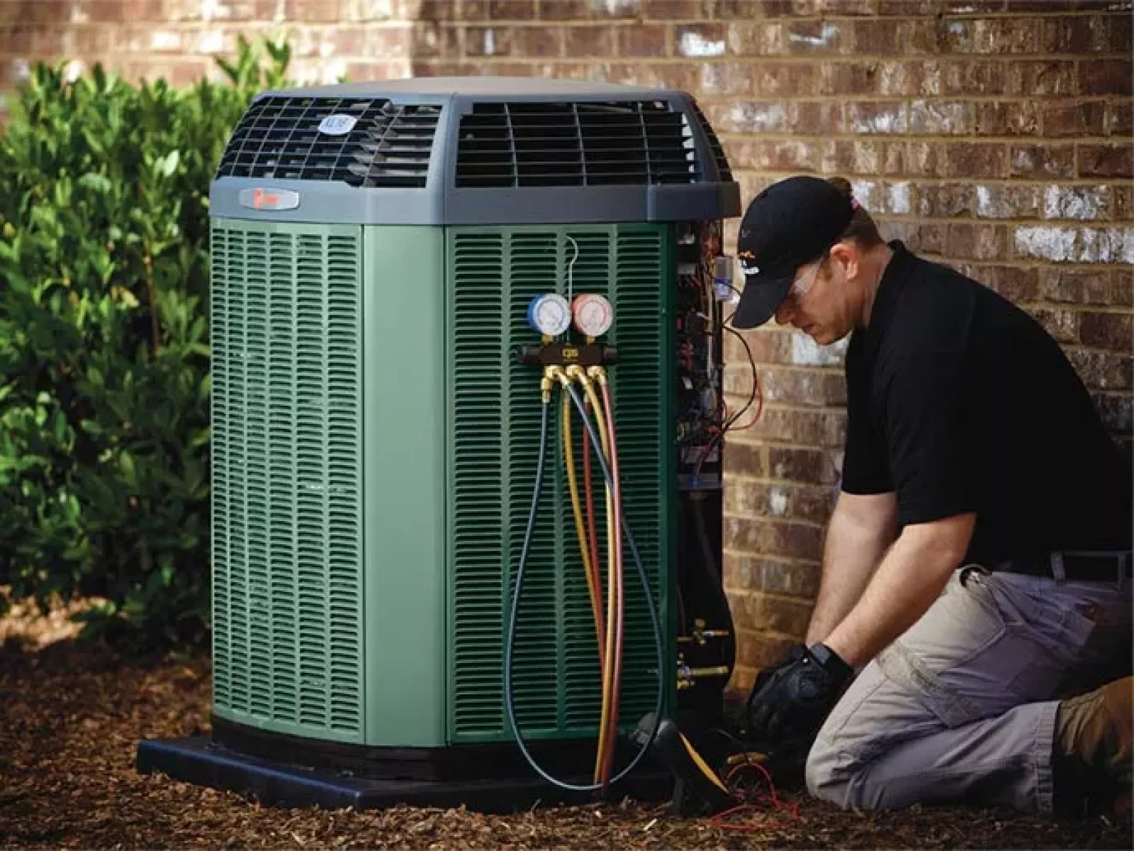 Technician servicing a green outdoor air conditioning unit next to a brick wall with gauges and wires attached