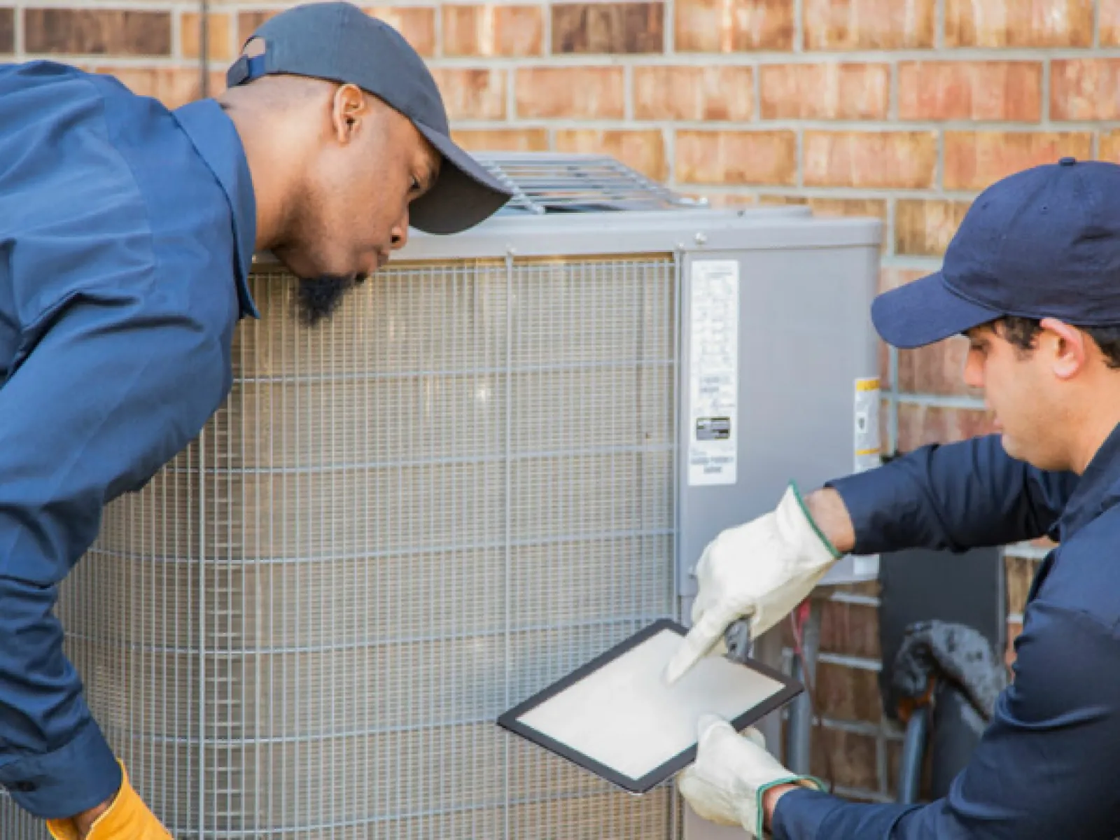 Two HVAC technicians inspecting and replacing an air filter on an outdoor air conditioning unit by a brick wall.