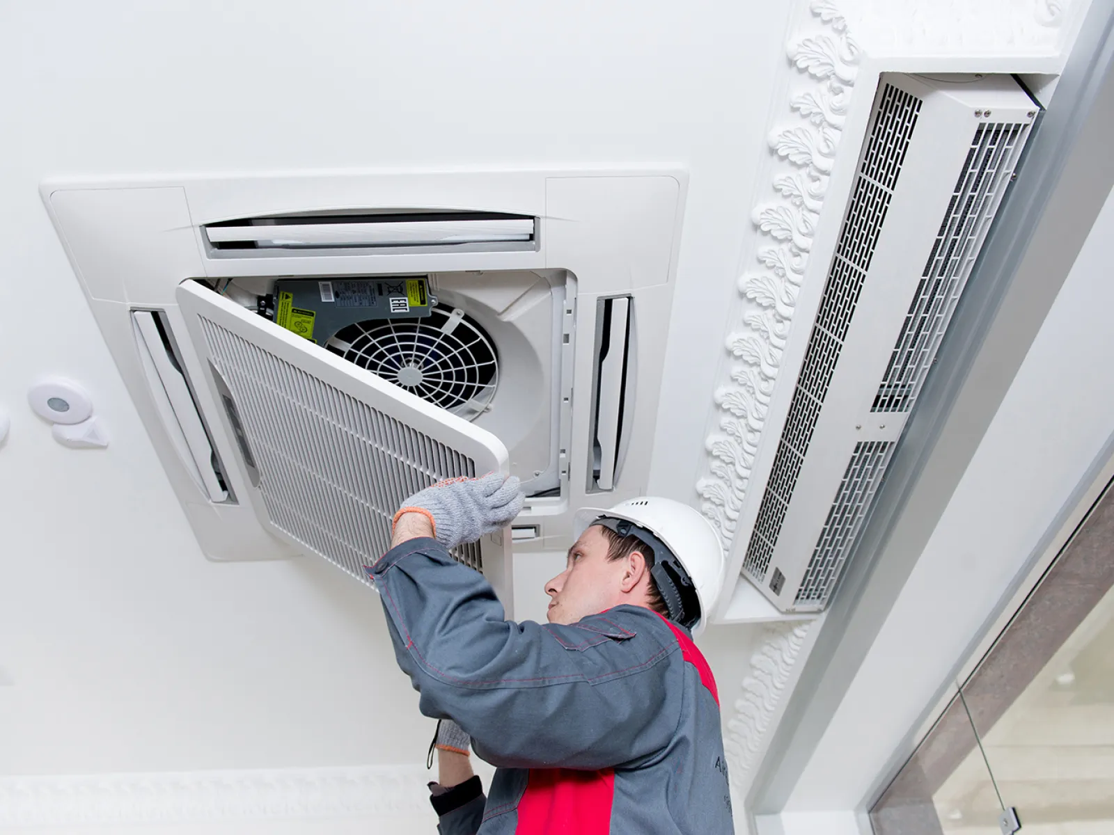 Technician wearing helmet and gloves servicing a ceiling-mounted air conditioning unit with the cover panel removed.