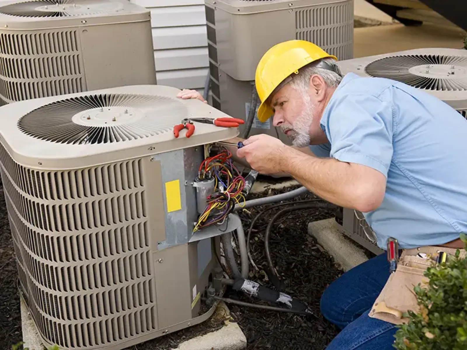 Technician in yellow hard hat repairing an outdoor air conditioning unit with tools and wiring exposed.