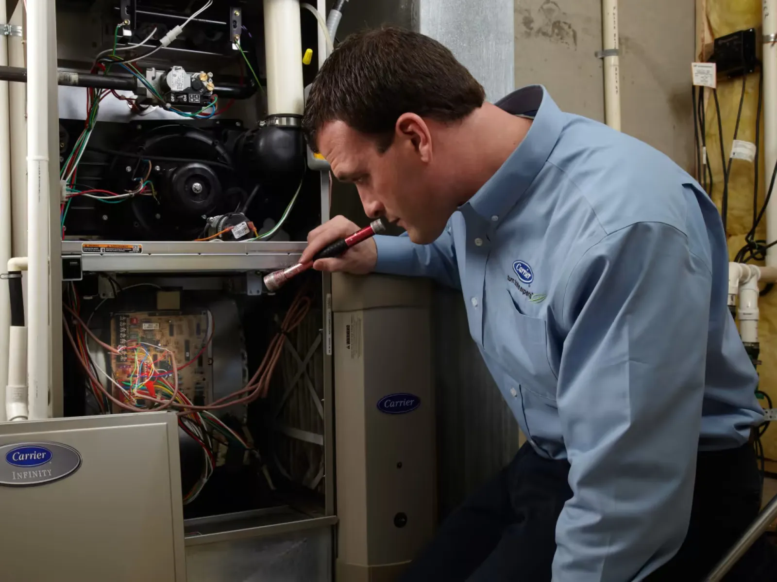 Technician inspecting and repairing a Carrier HVAC system with a flashlight in a residential setting.