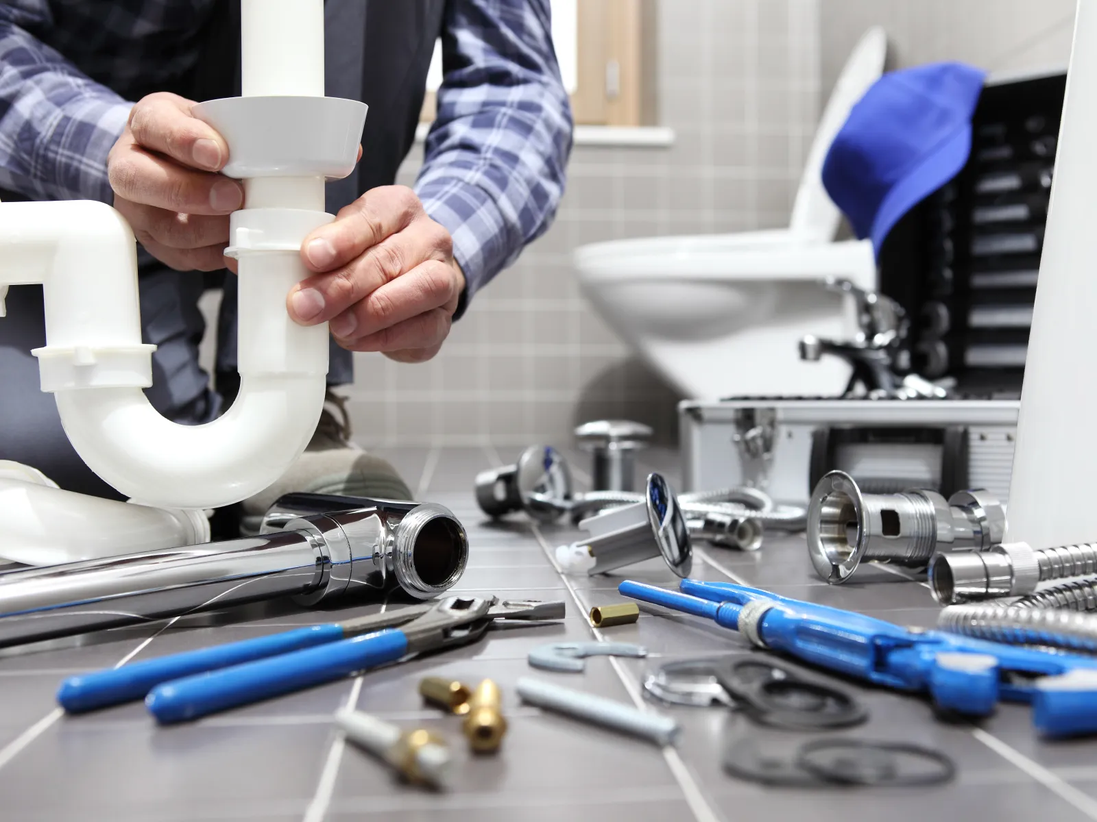 Plumber fitting a white PVC pipe under a bathroom sink with various plumbing tools scattered on the floor.