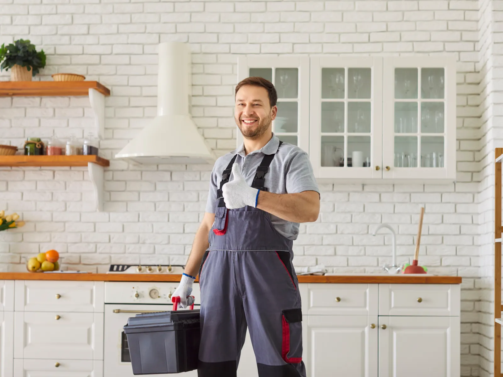 Smiling handyman in overalls giving thumbs up while holding a toolbox in a modern white kitchen.