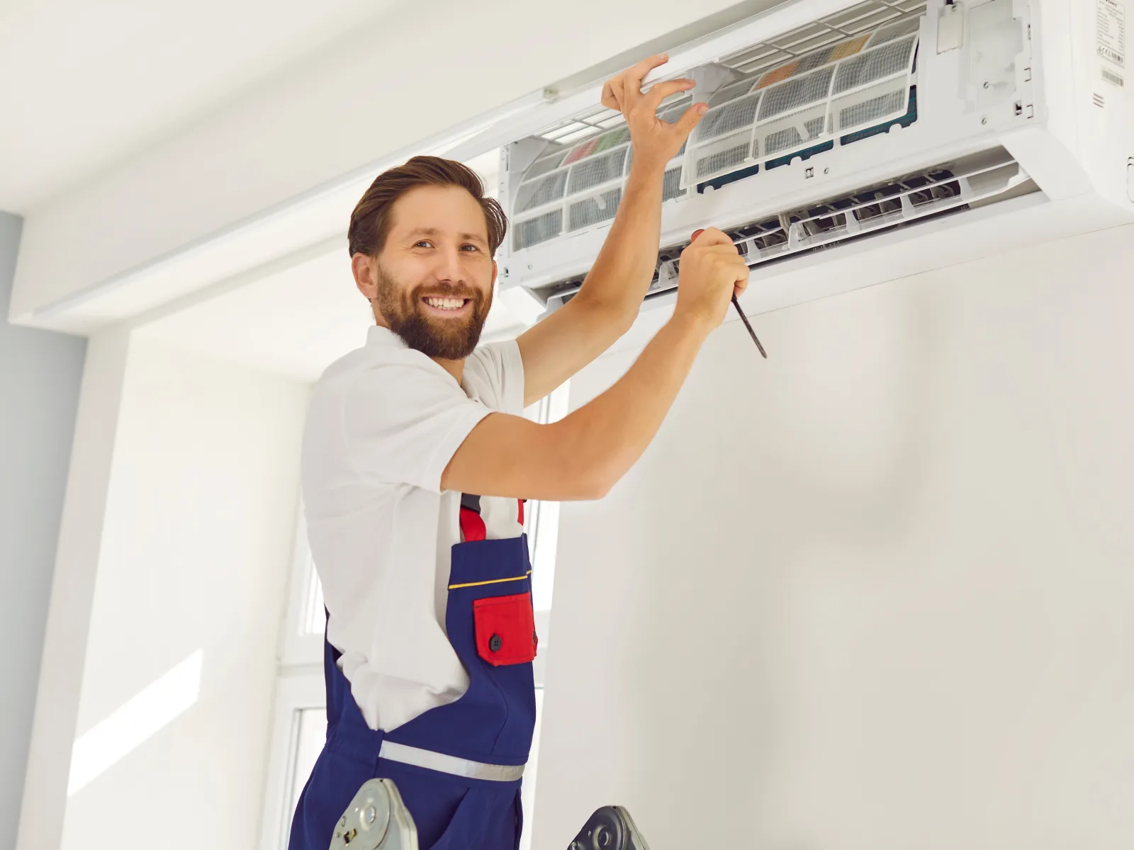Technician smiling while repairing a wall-mounted air conditioner in a bright room using a screwdriver.
