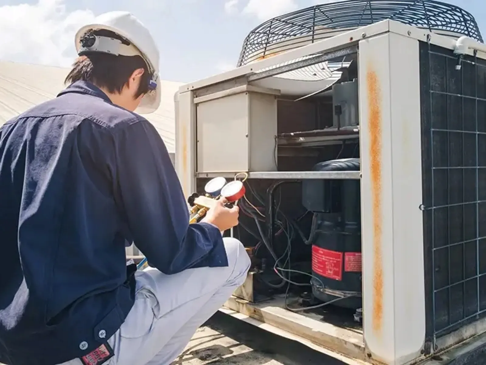 Technician in helmet inspecting and repairing outdoor air conditioning unit with gauges under clear sky.