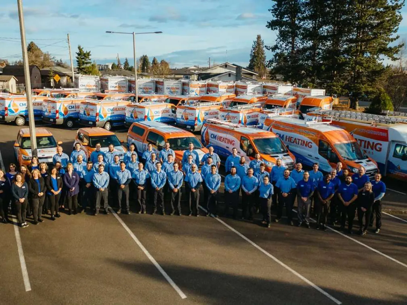 Large team in blue uniforms standing in front of a fleet of CM Heating branded vans in a parking lot on a sunny day