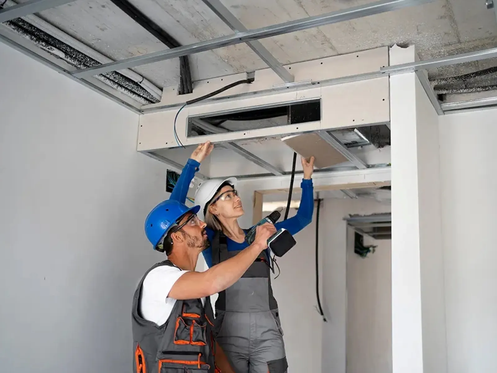 Two construction workers installing ceiling panels inside a building under renovation with safety helmets and tools.