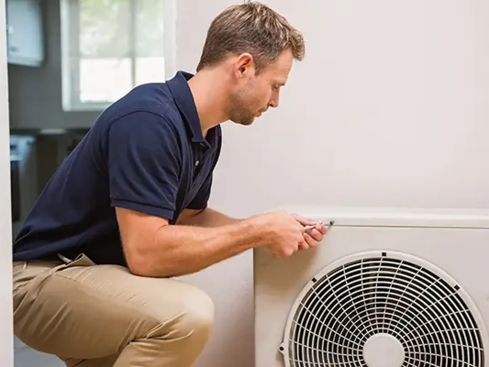 Technician in navy shirt and khaki pants repairing an indoor HVAC unit with a screwdriver in a bright room.