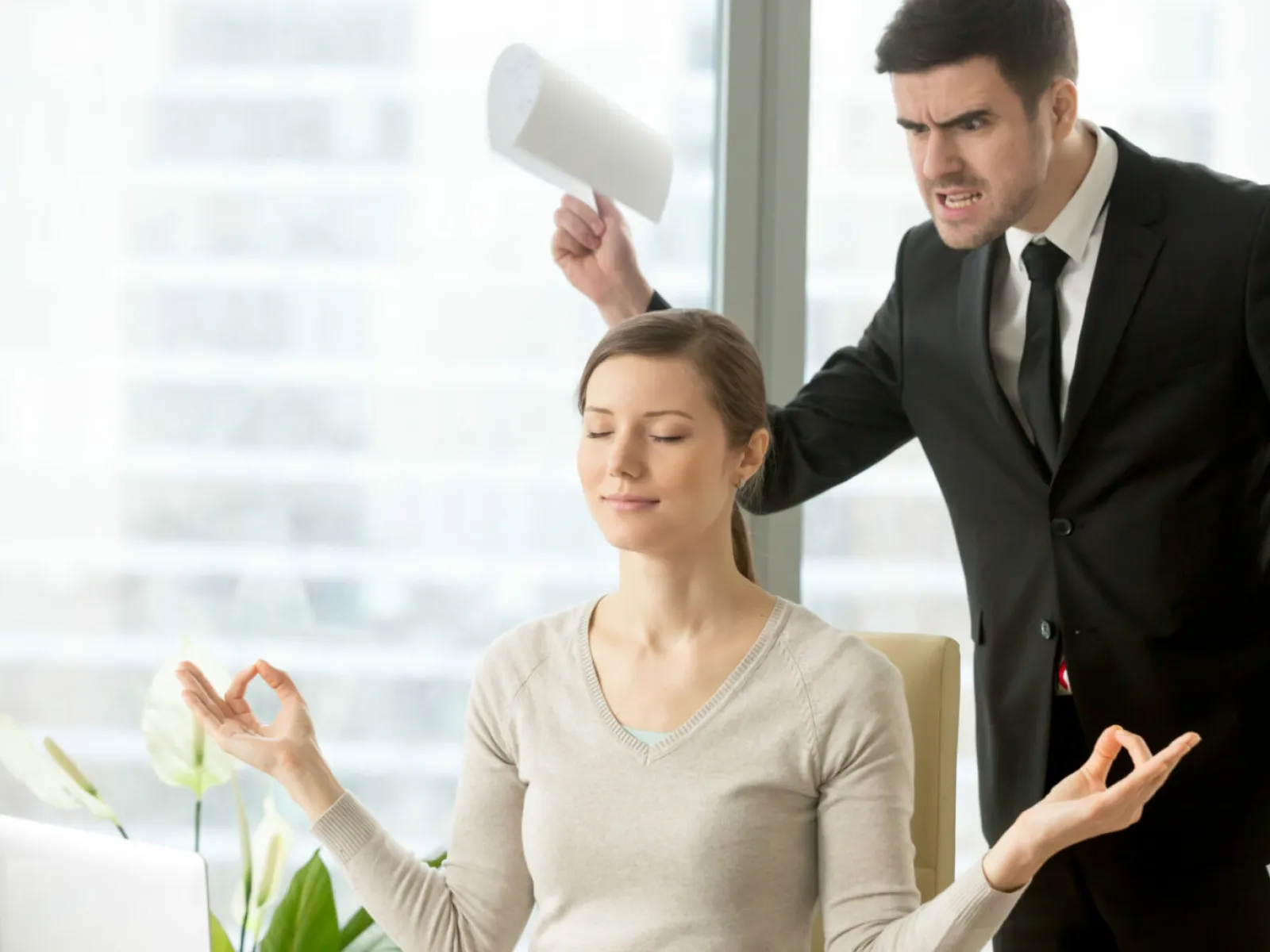 Calm woman meditating in office while angry man yells and raises paper in frustration behind her.
