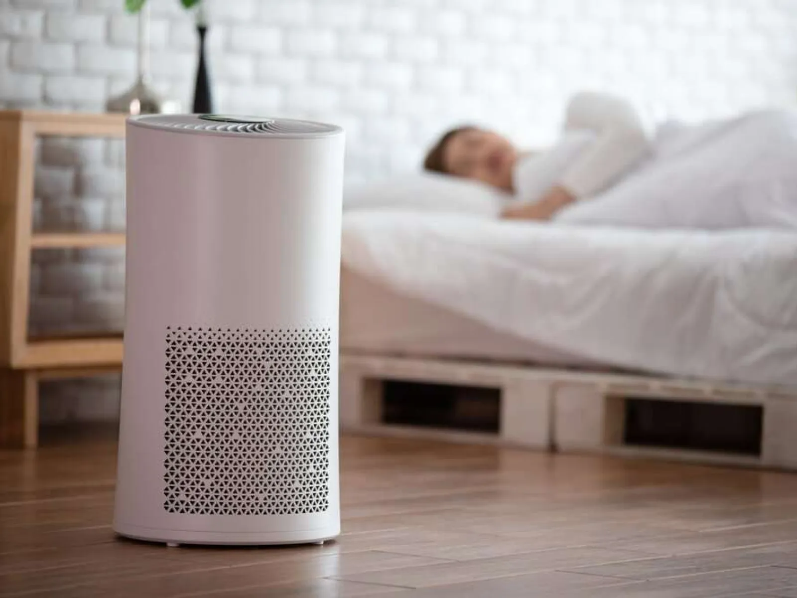 Modern white air purifier on wooden floor with sleeping person in bed in background for clean air concept