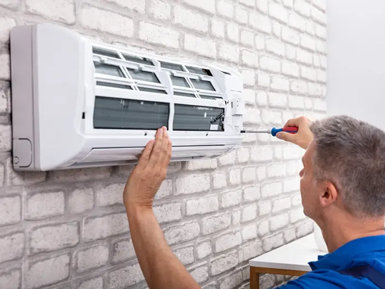 Technician in blue shirt repairing or maintaining a white wall-mounted air conditioner with a screwdriver indoors.