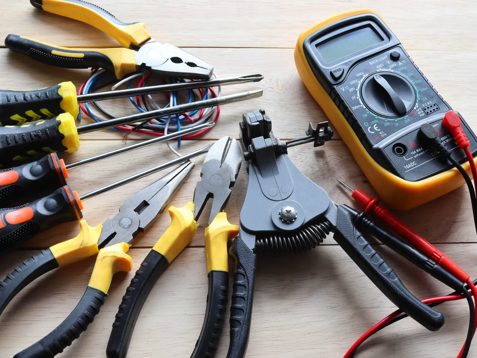 Set of electrical tools including pliers, screwdrivers, wire strippers, and a digital multimeter on wooden surface
