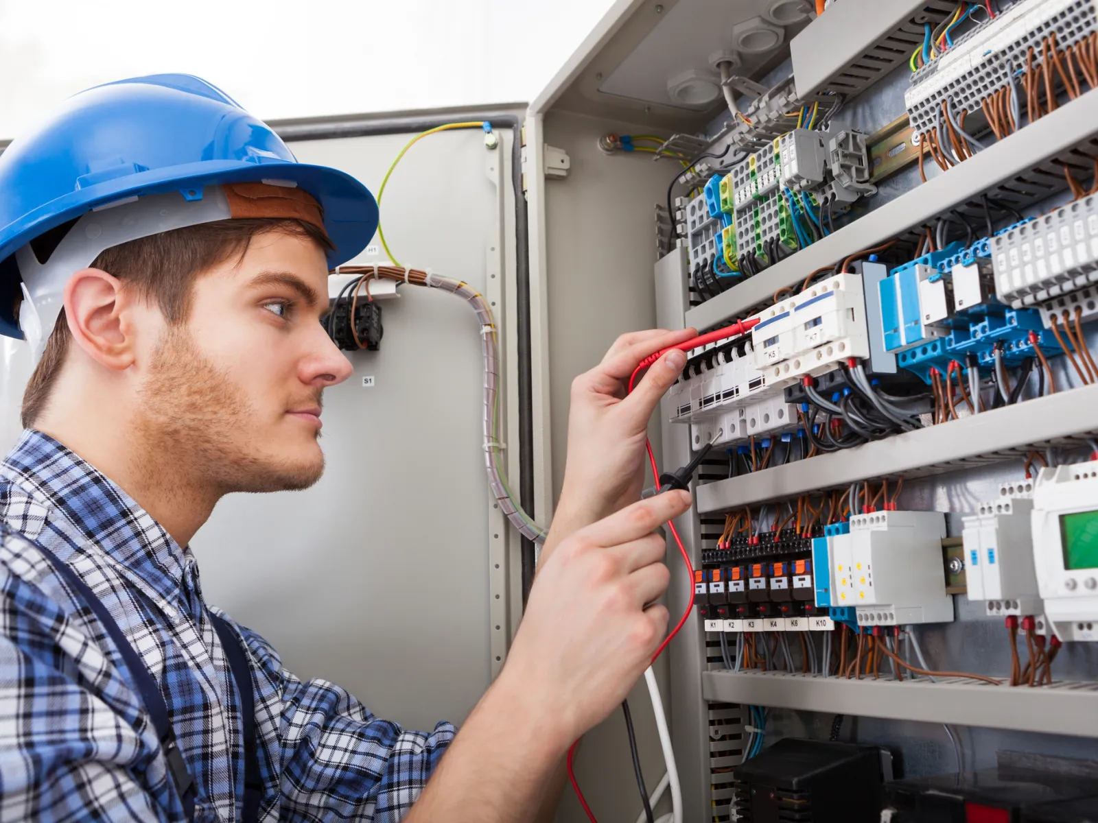 Electrician in blue hard hat working on electrical panel with wiring and circuit breakers indoors.