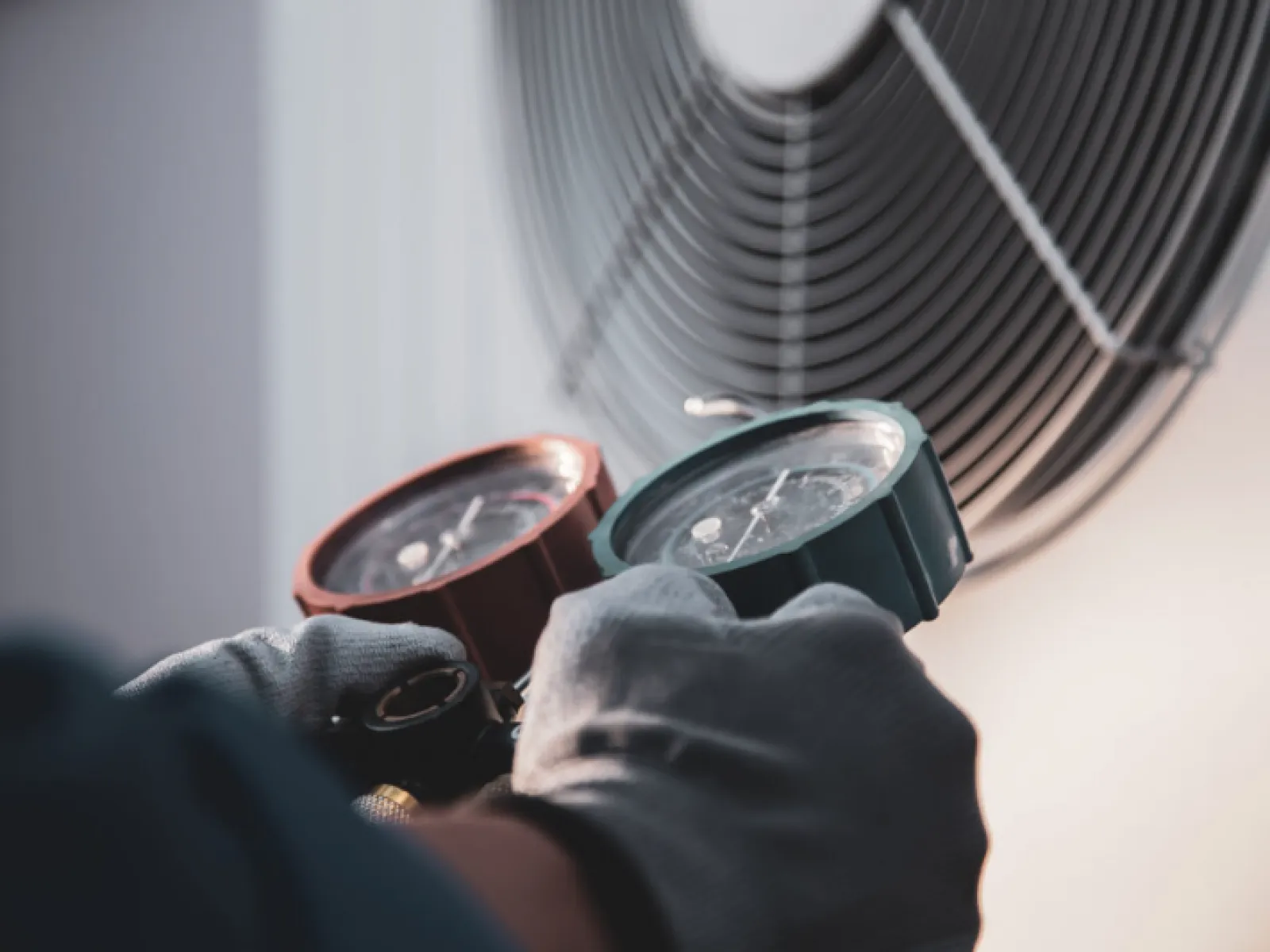 Technician in gloves measuring air conditioning pressure using HVAC gauges near a cooling fan.
