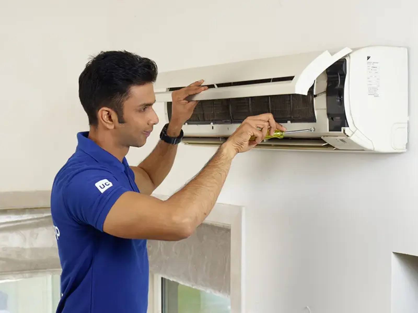 Technician in blue shirt repairing a wall-mounted air conditioner with a screwdriver indoors.