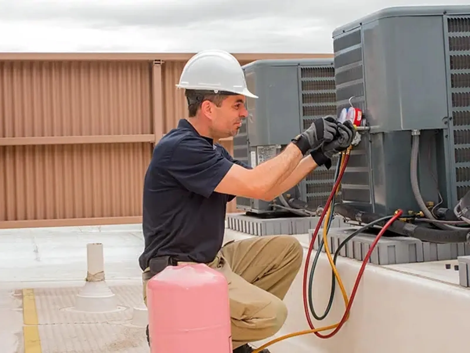Technician in safety helmet and gloves working on rooftop HVAC unit with gauges and hoses for maintenance.