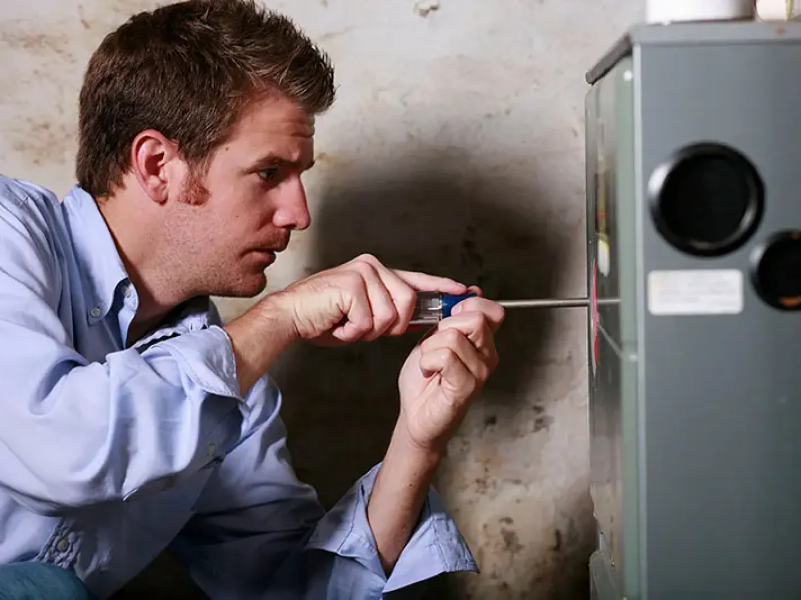 Man in blue shirt using screwdriver to repair or adjust furnace or HVAC unit in a basement.