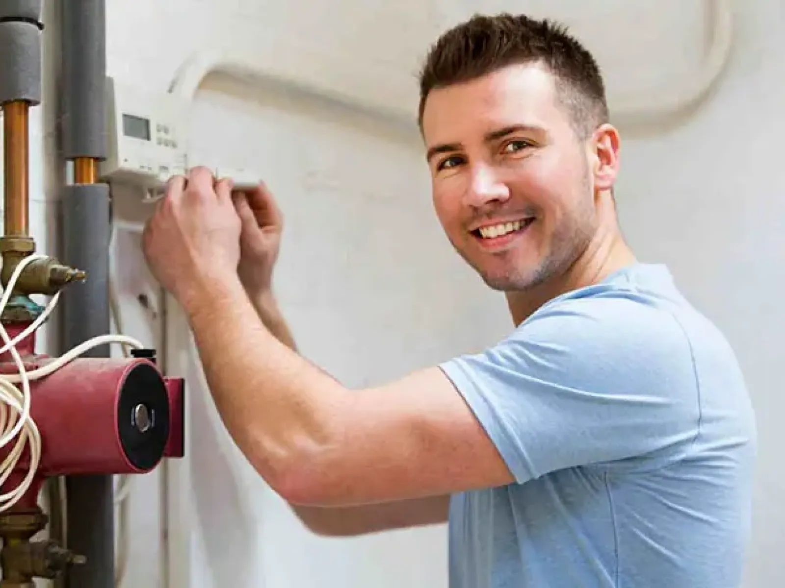 Smiling man adjusting a thermostat on a wall near home plumbing pipes and a boiler unit.