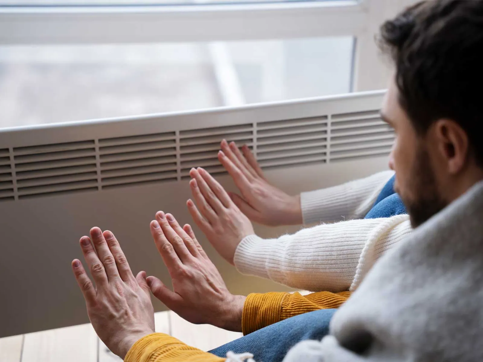 People warming their hands near a radiator heater indoors on a cold day