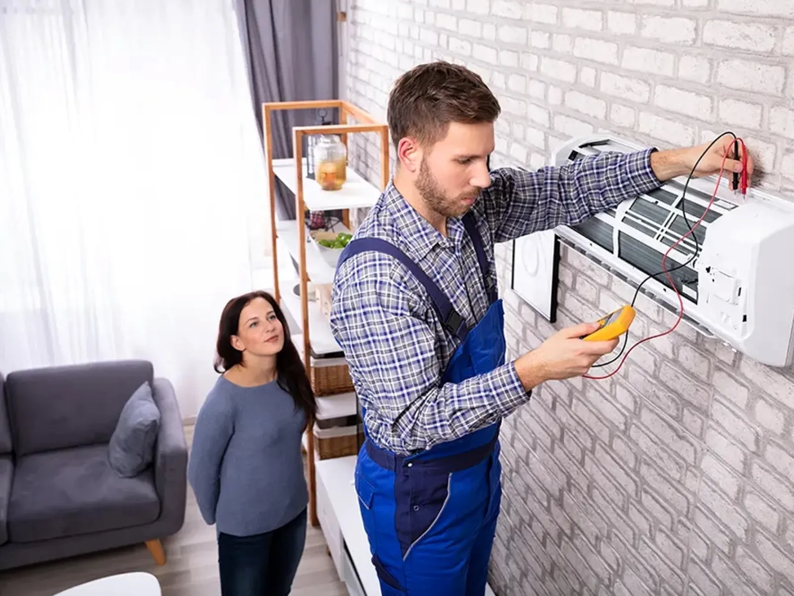 Technician in blue overalls repairing wall-mounted air conditioner while woman watches in living room.