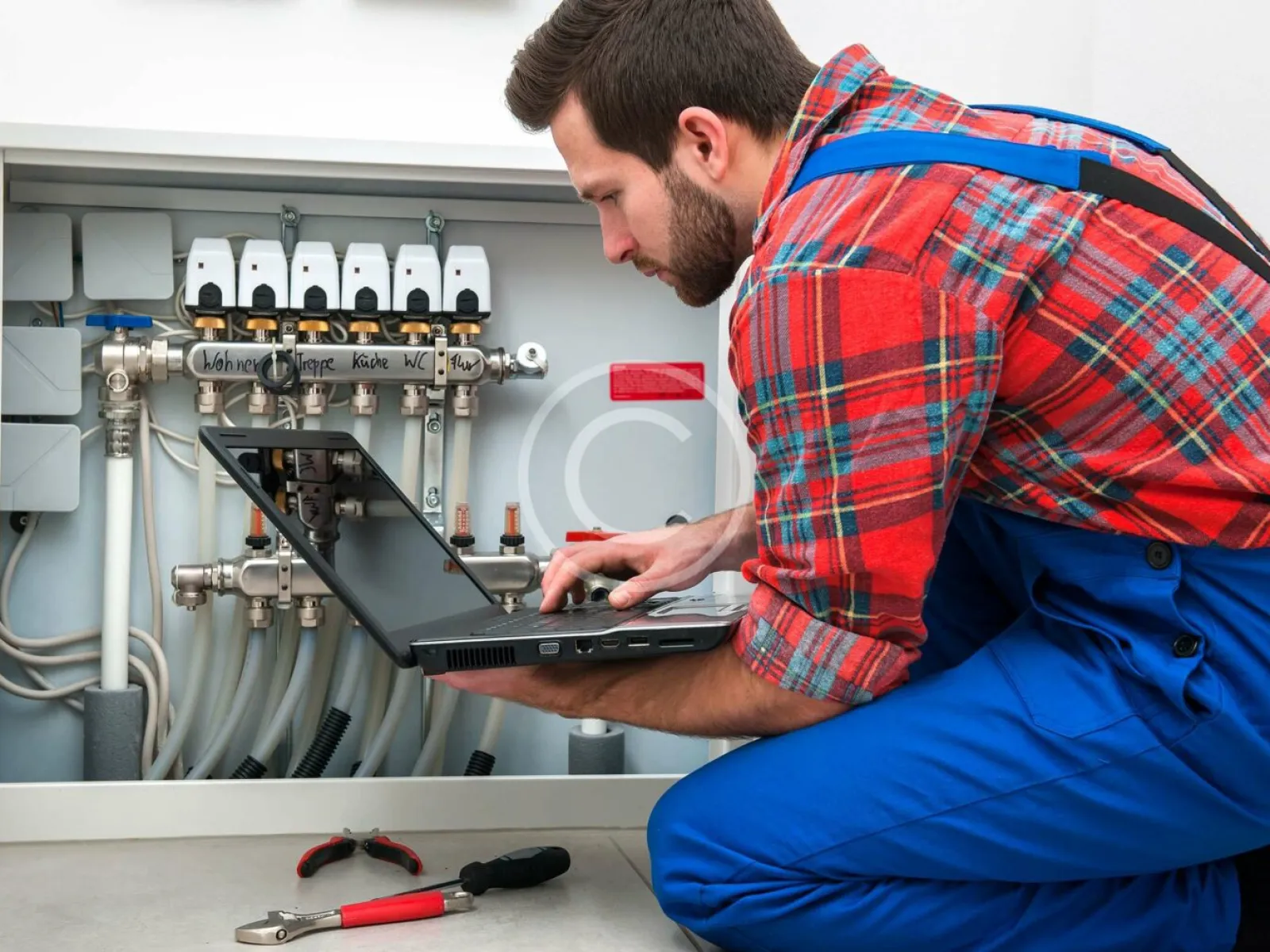 Technician in blue overalls using laptop to inspect plumbing system under sink with tools nearby.