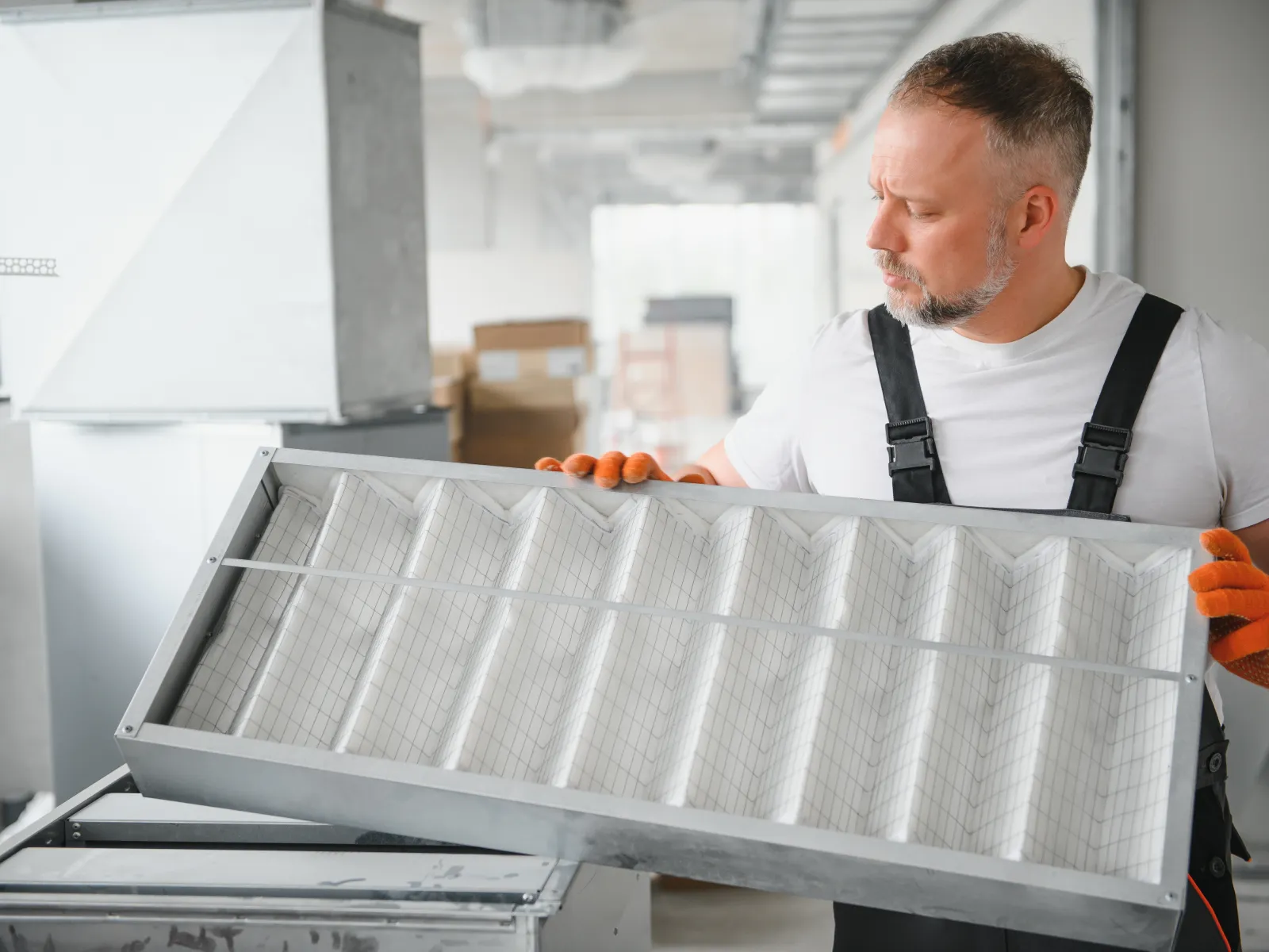 Technician in white shirt and orange gloves inspecting a large rectangular HVAC air filter in an industrial setting