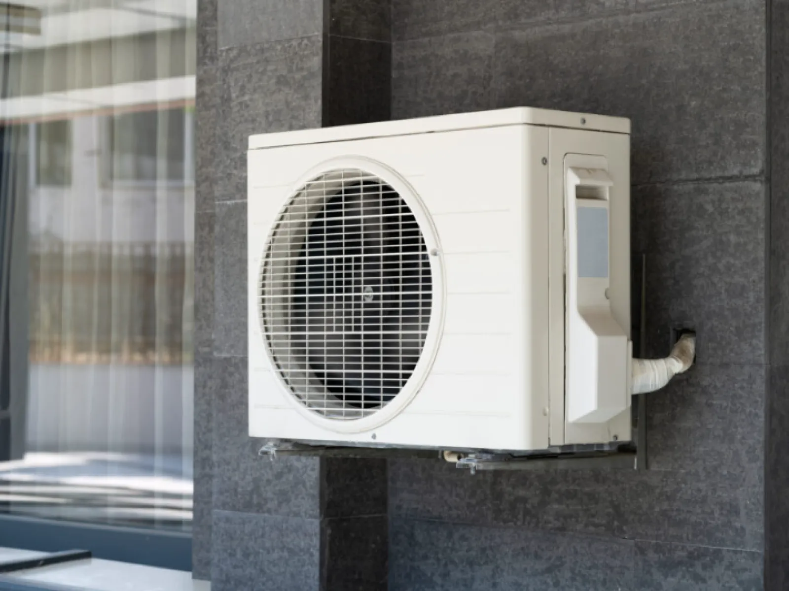 White air conditioning unit mounted on dark gray exterior wall near window of modern building.