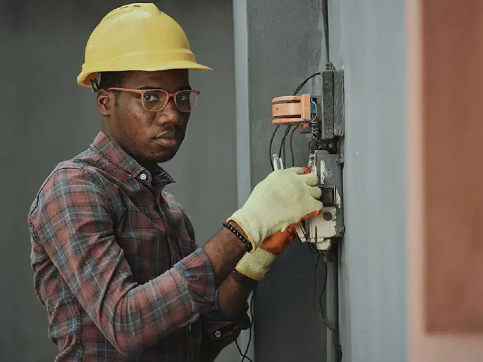 Electrician wearing a yellow hard hat and gloves working on an electrical switch on a gray wall.
