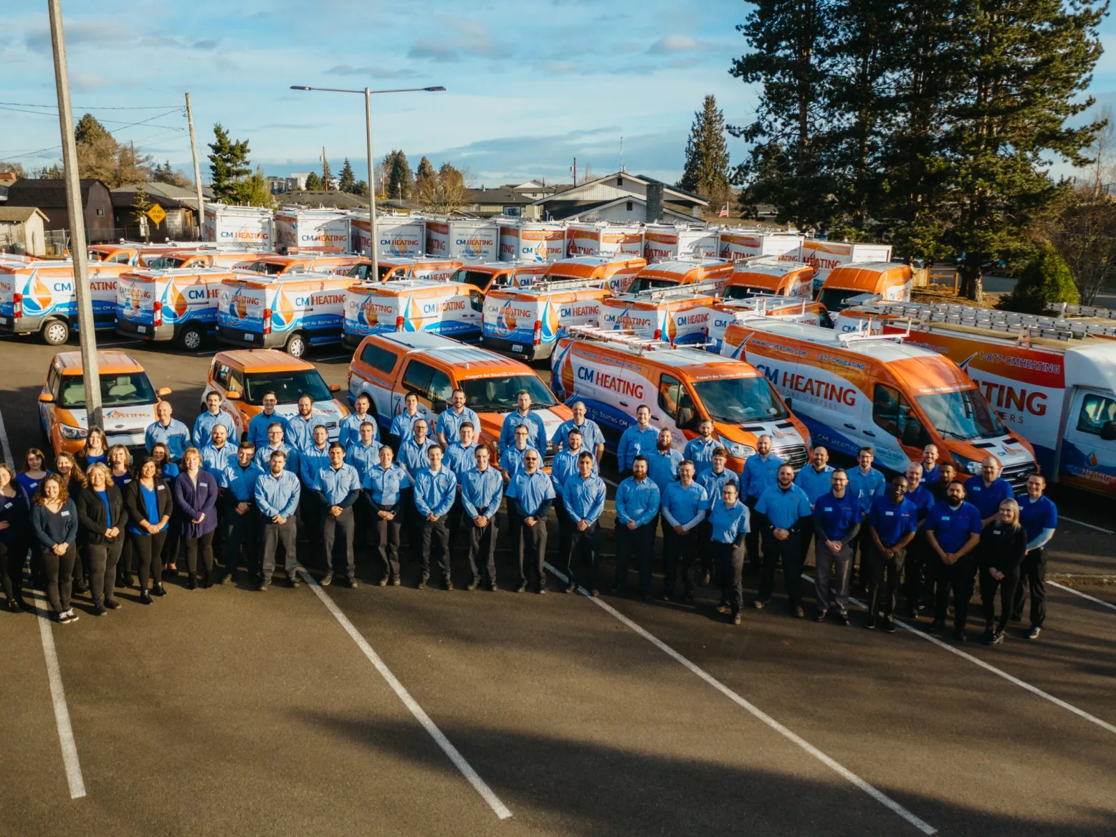 Group of CM Heating employees standing in front of company vans and trucks in parking lot under clear sky.