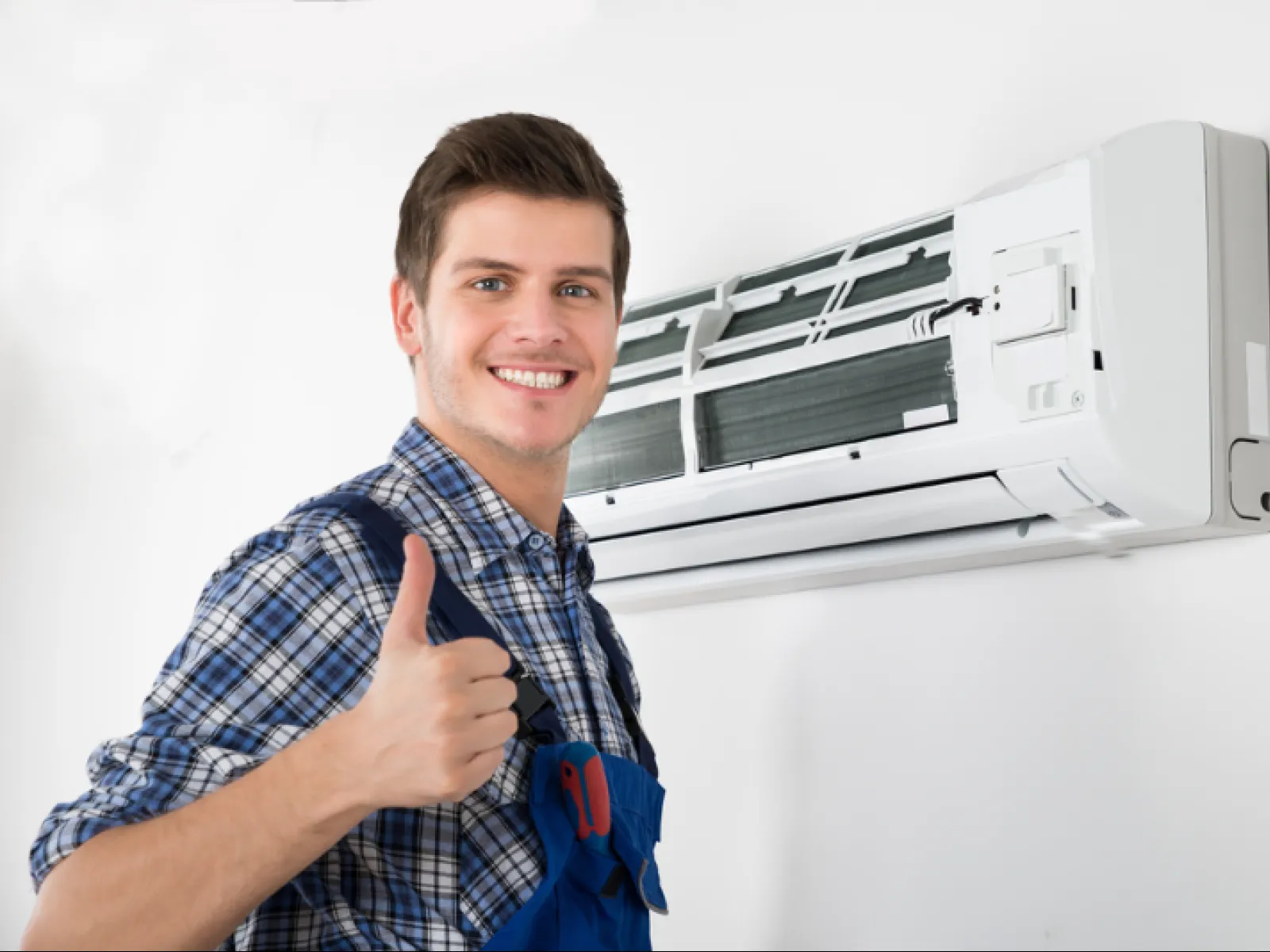 Technician in blue overalls gives thumbs up next to a wall-mounted air conditioner unit during service.