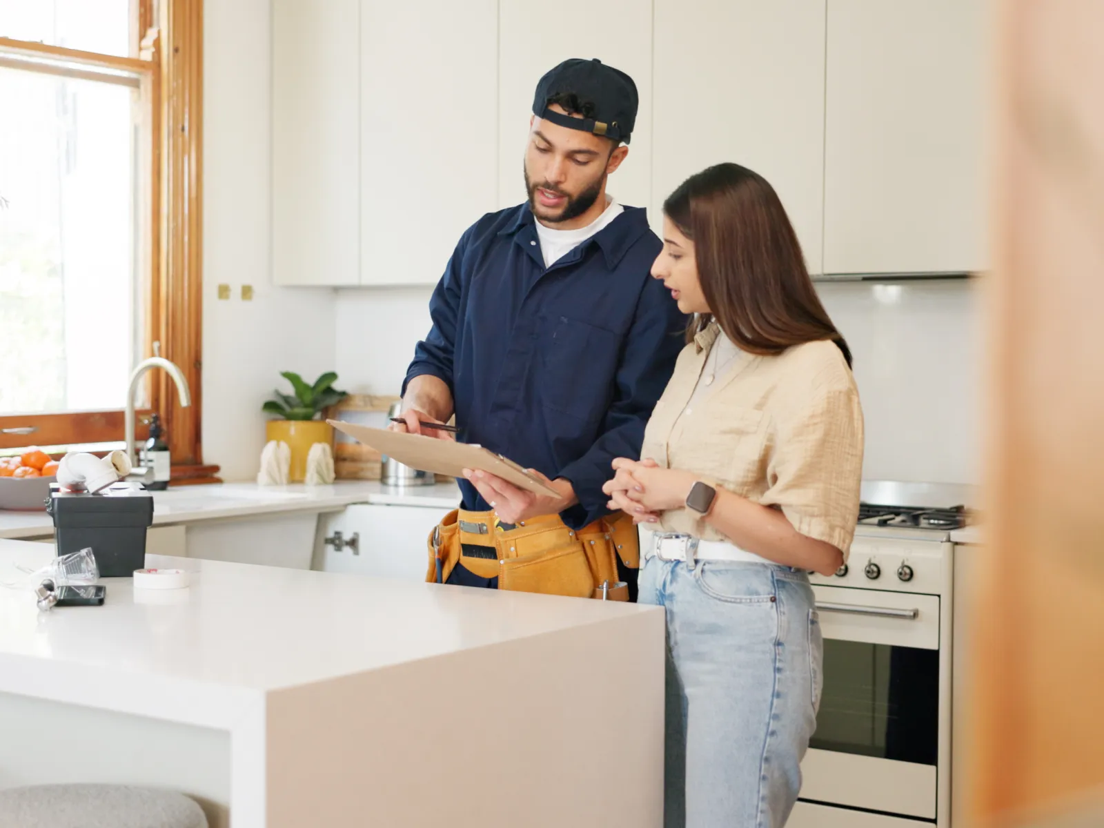 Man in work uniform explaining repairs to woman in modern kitchen with white countertops and natural light