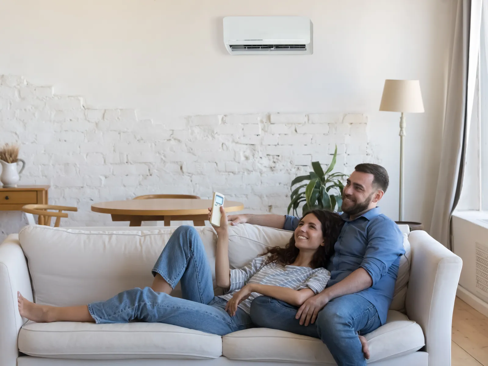 Couple relaxing on white couch using remote control in bright living room with air conditioner.