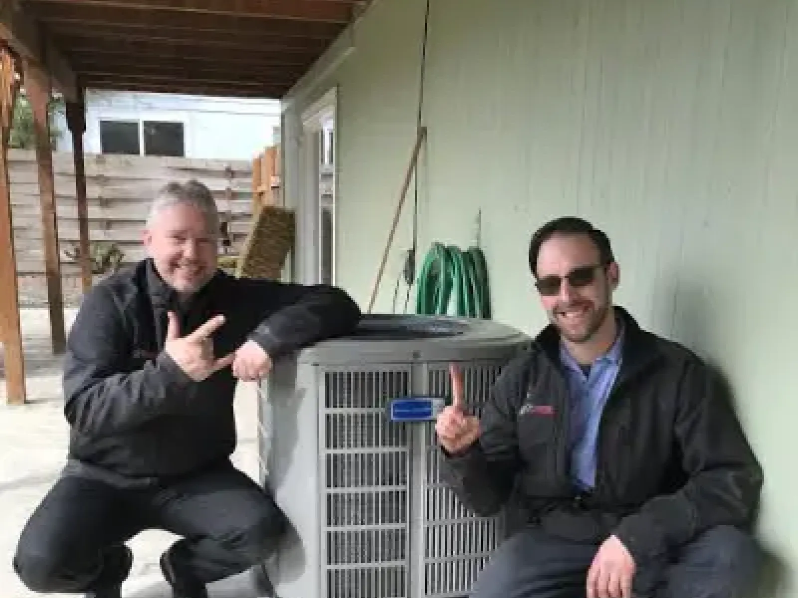 Two men in black jackets crouch beside a newly installed outdoor air conditioning unit smiling and gesturing.
