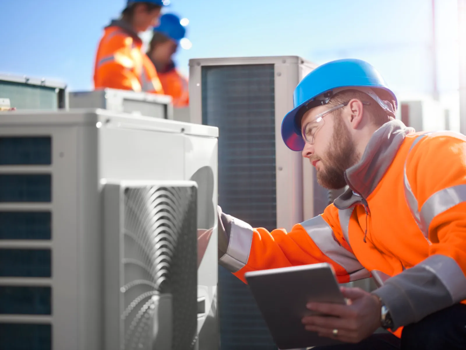 Technician in orange safety jacket and blue helmet inspecting HVAC unit with a tablet on a sunny day.