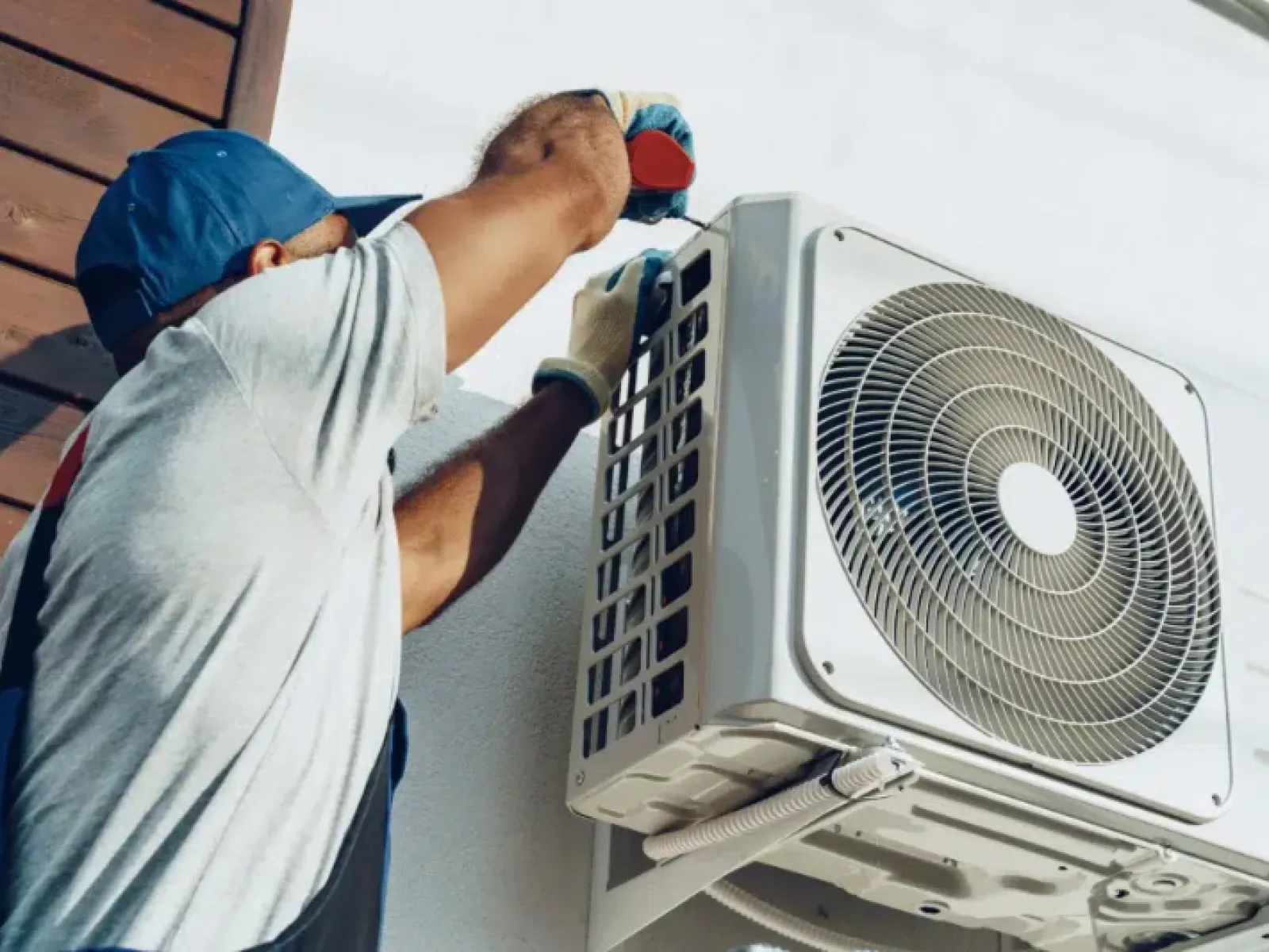 Technician in blue cap and gloves installing an outdoor air conditioning unit on a white wall.