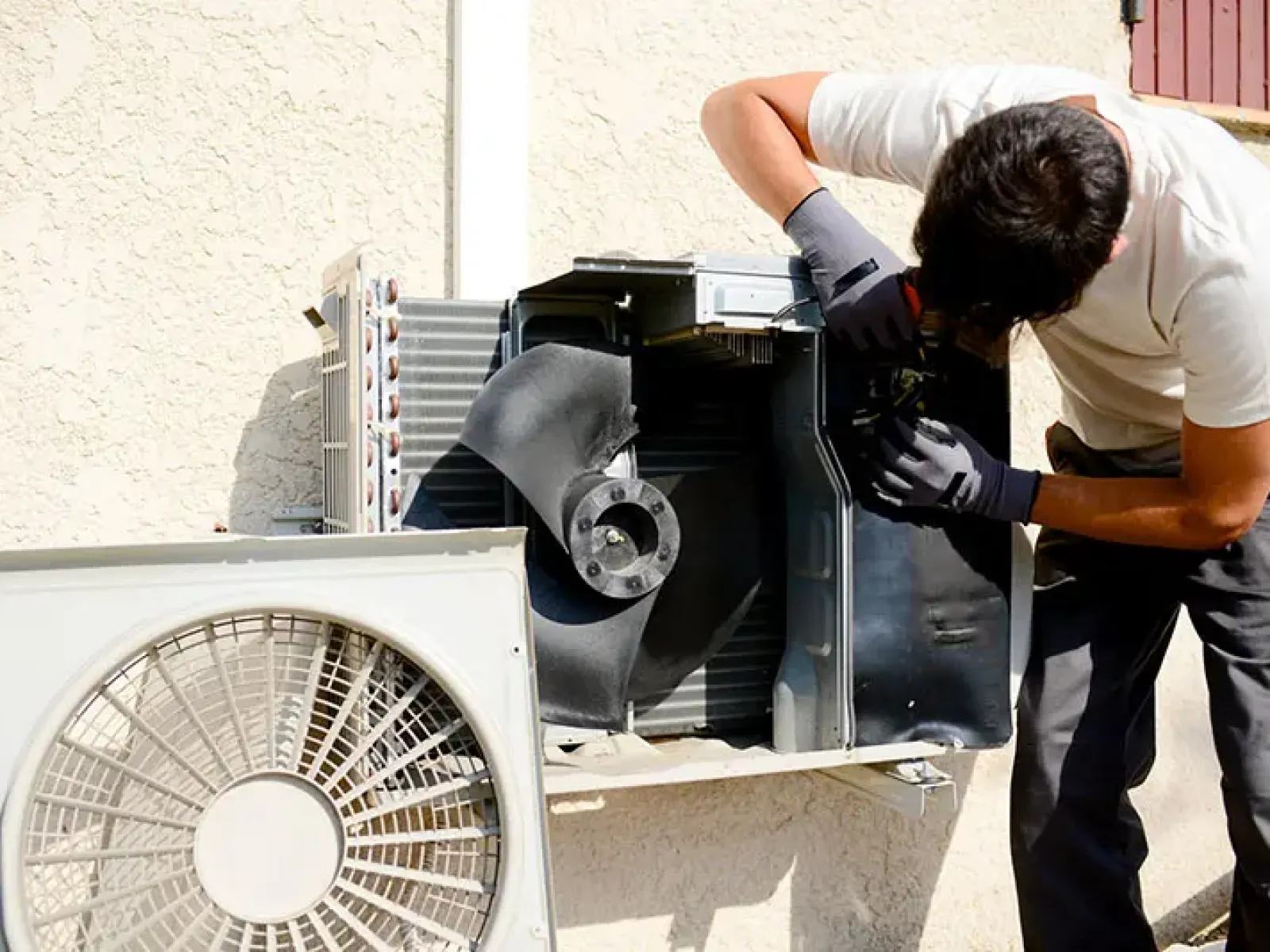 Technician repairing an outdoor air conditioning unit mounted on a beige wall under sunlight.