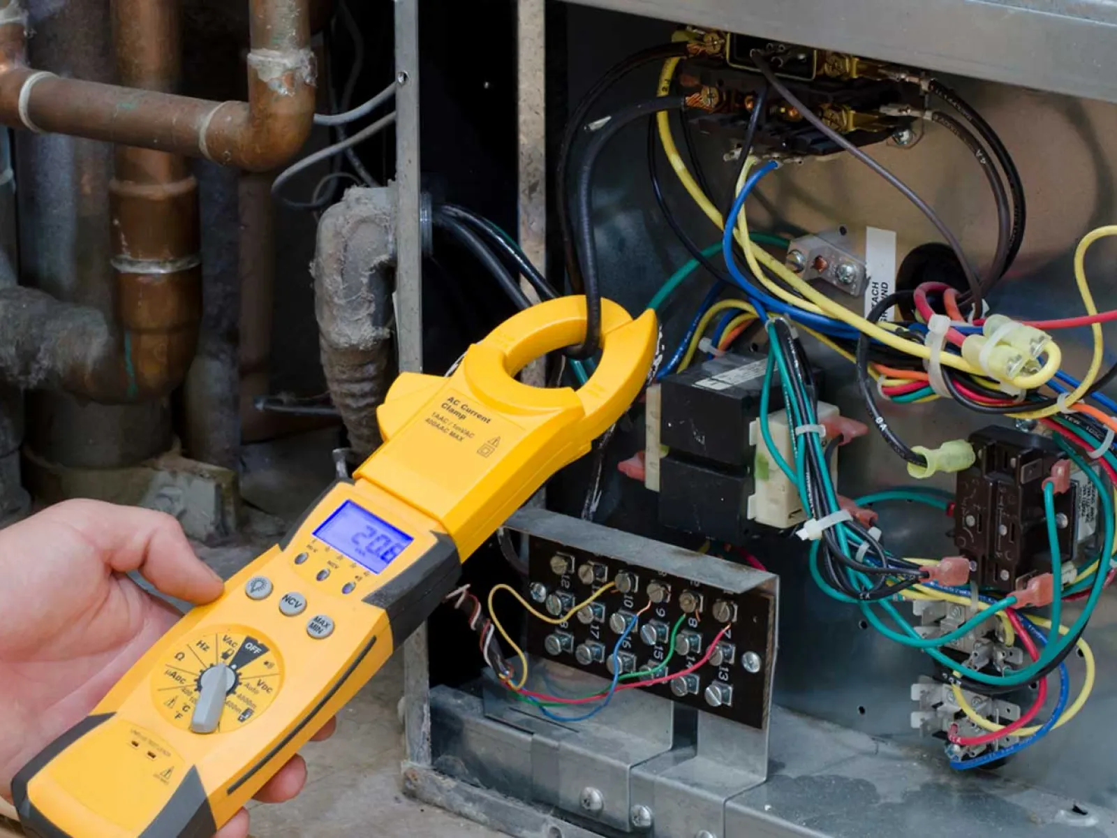 Hand holding a yellow clamp meter measuring electrical current in a complex wiring panel with multicolored wires.