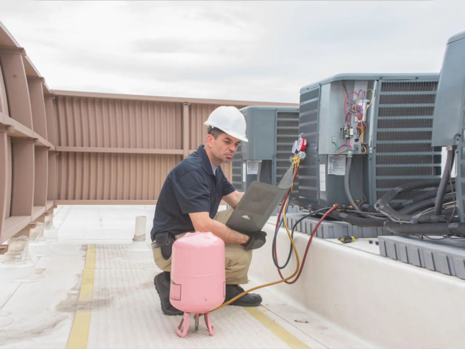 Technician in hard hat using laptop to inspect rooftop HVAC units with tools and gauges nearby
