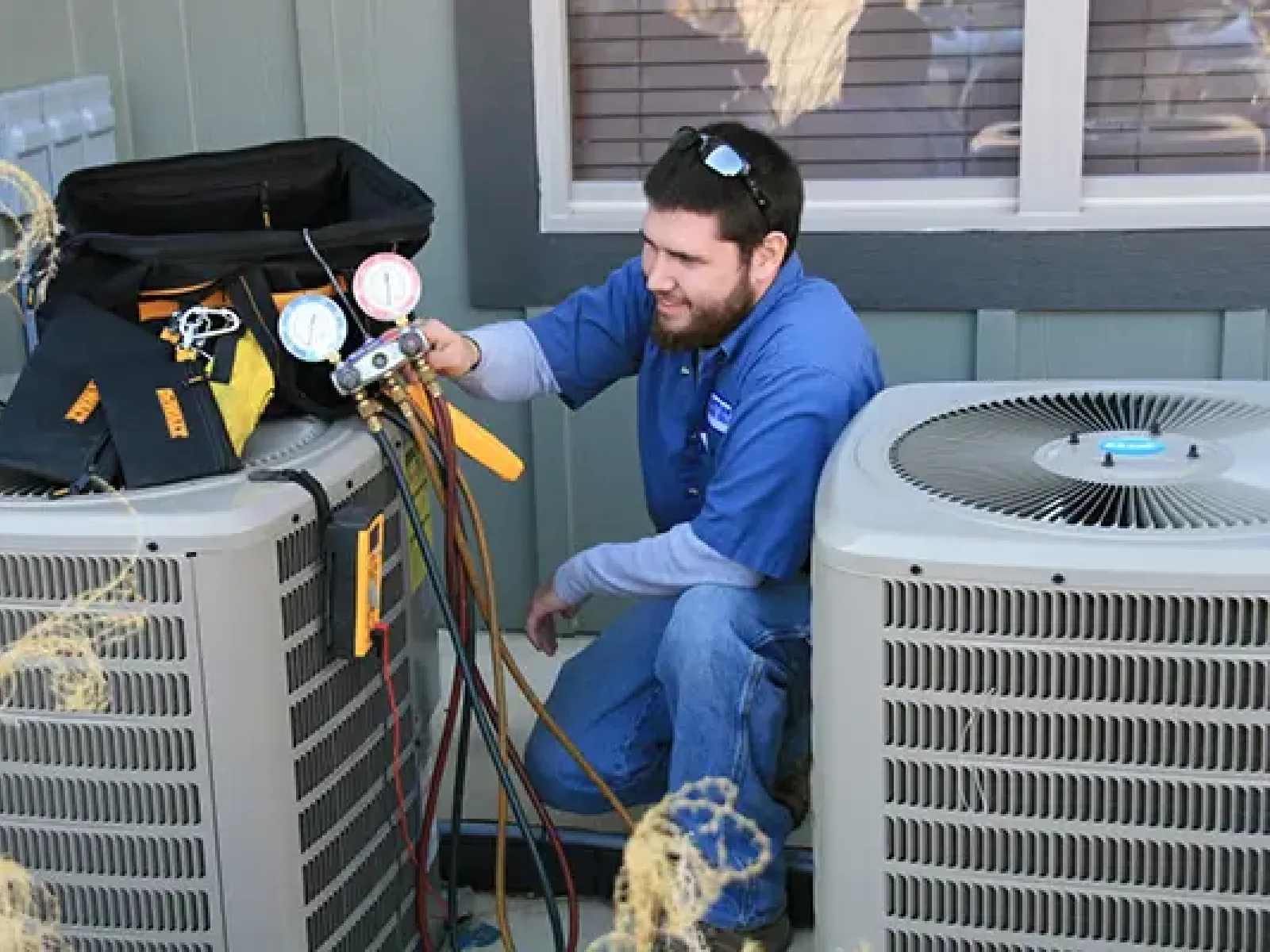 Technician in blue uniform servicing air conditioning units with gauges and tools outside a house.