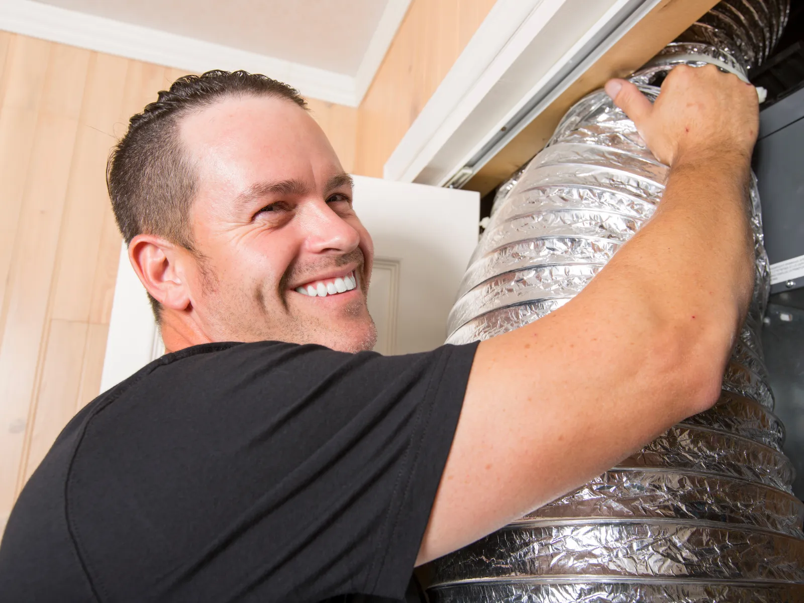 Smiling technician installing or repairing flexible HVAC ducting inside a residential home with wooden panel walls.