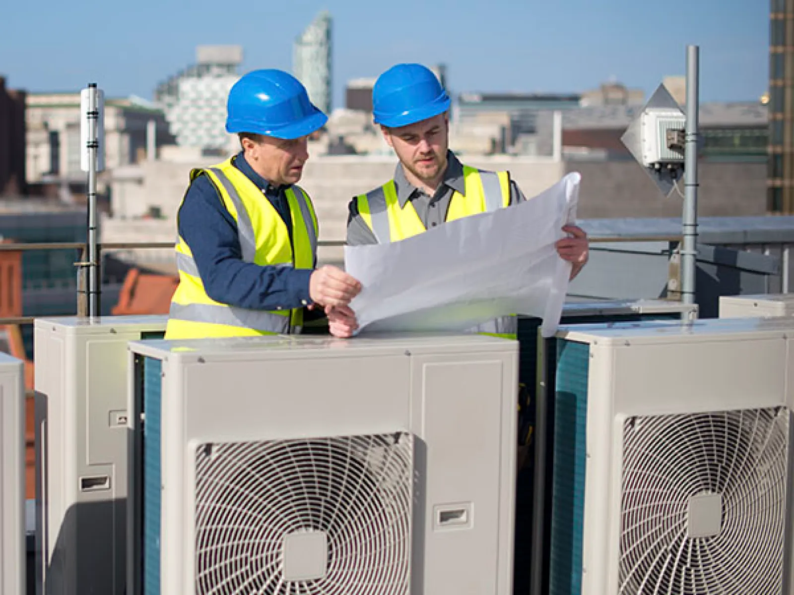 Two construction workers in hard hats and safety vests reviewing plans on a rooftop with HVAC units and city buildings.