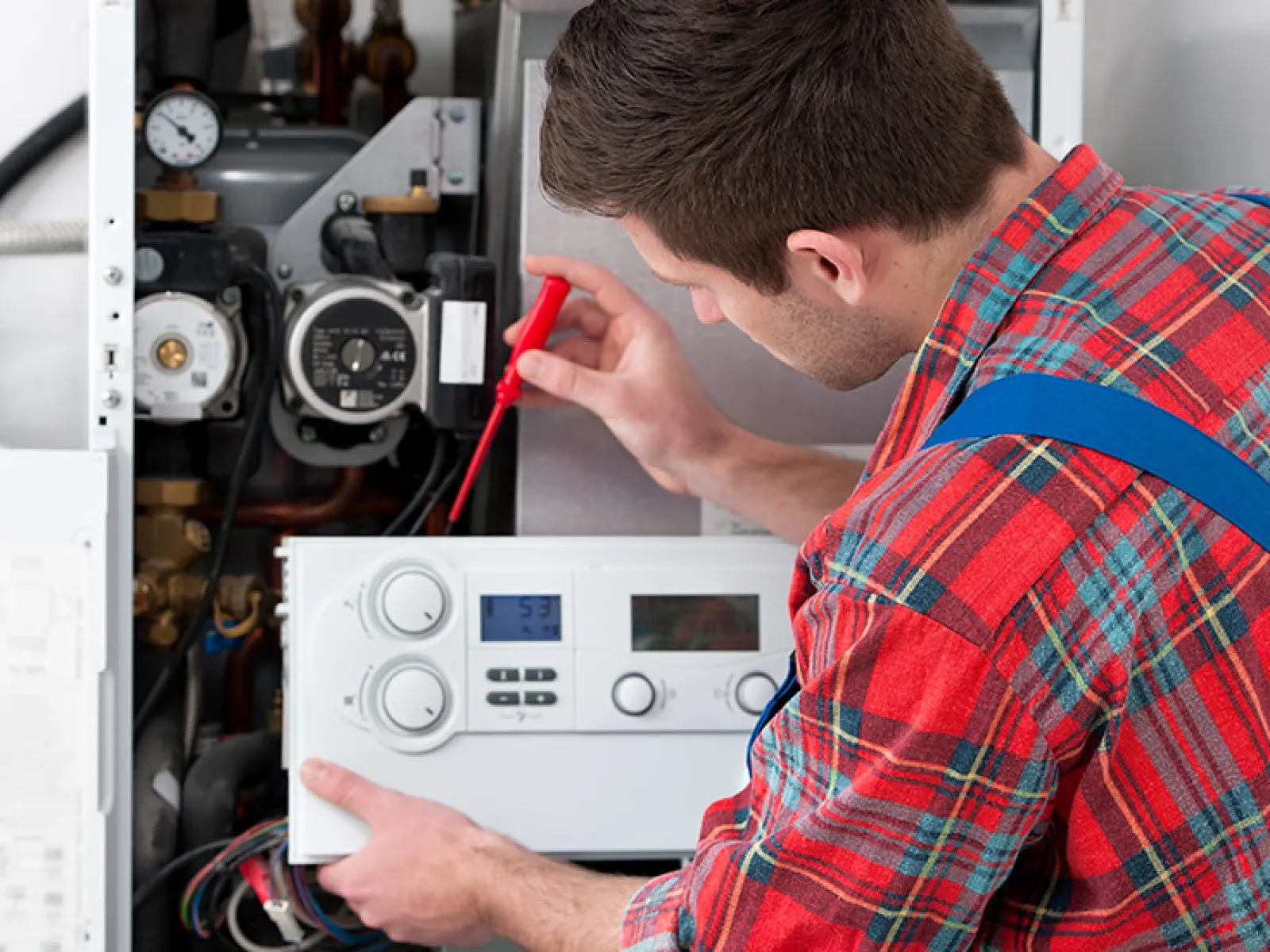 Technician in plaid shirt repairing or inspecting a boiler or heating system with a screwdriver.