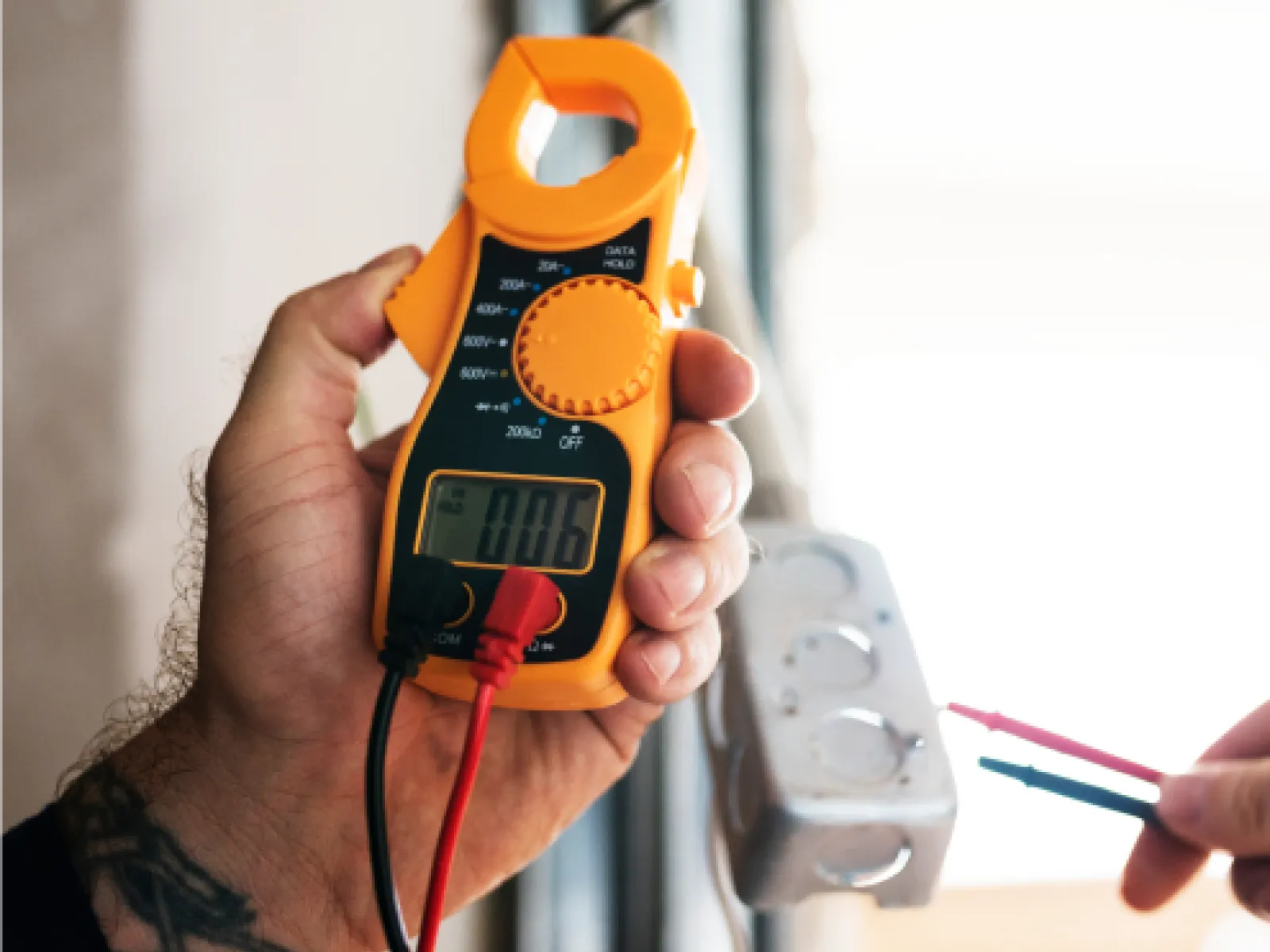 Close-up of hands using an orange digital clamp meter to measure electrical current on a circuit box.