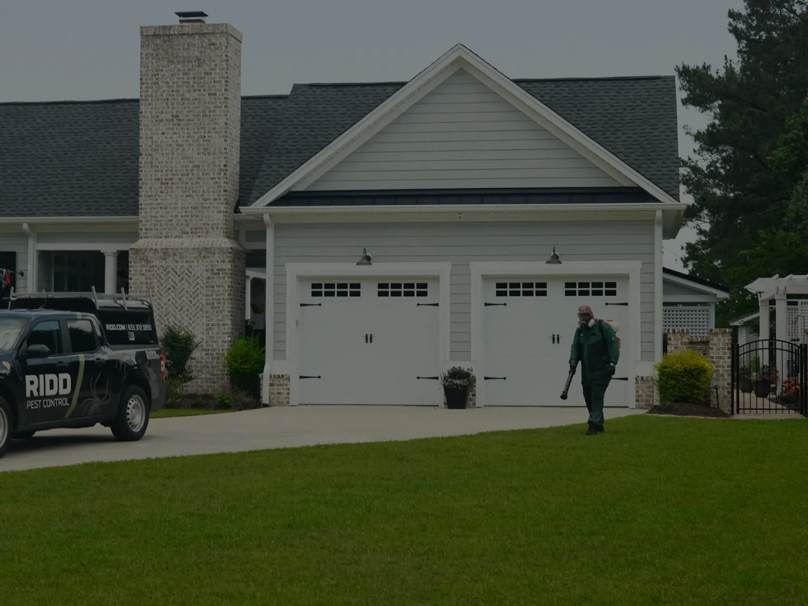 Pest control technician inspecting lawn near black RIDD Pest Control truck outside suburban white house.