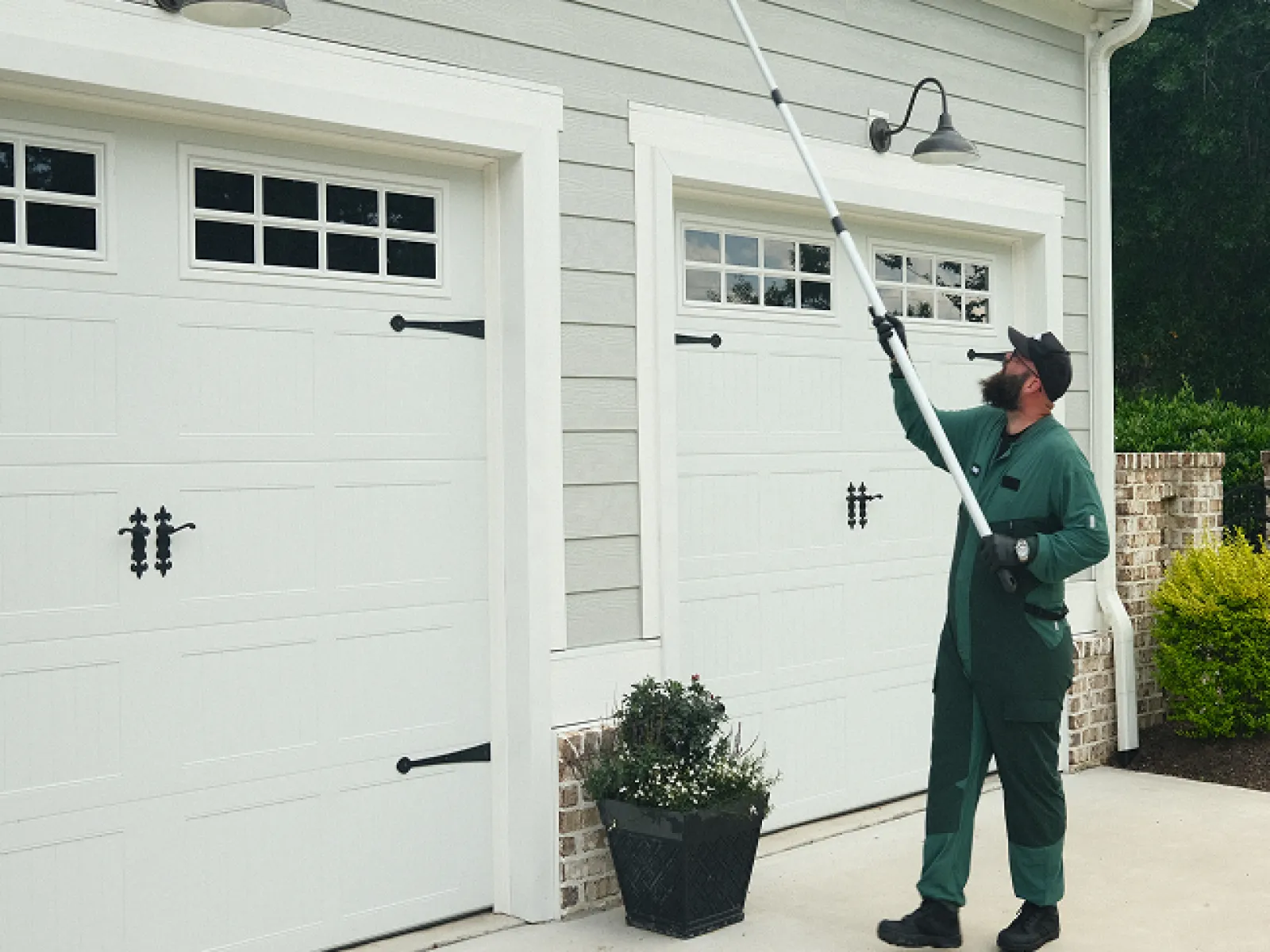 Man using long pole to clean exterior wall light fixtures on gray house with white garage doors.