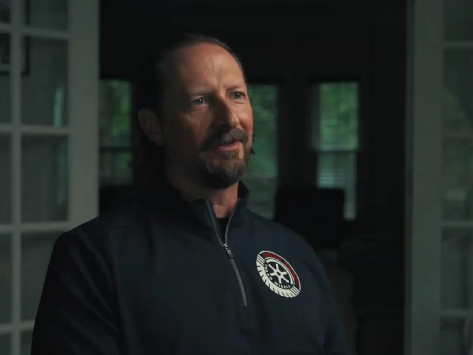 Man with beard wearing dark jacket with emblem, sitting indoors near glass doors in dimly lit room.