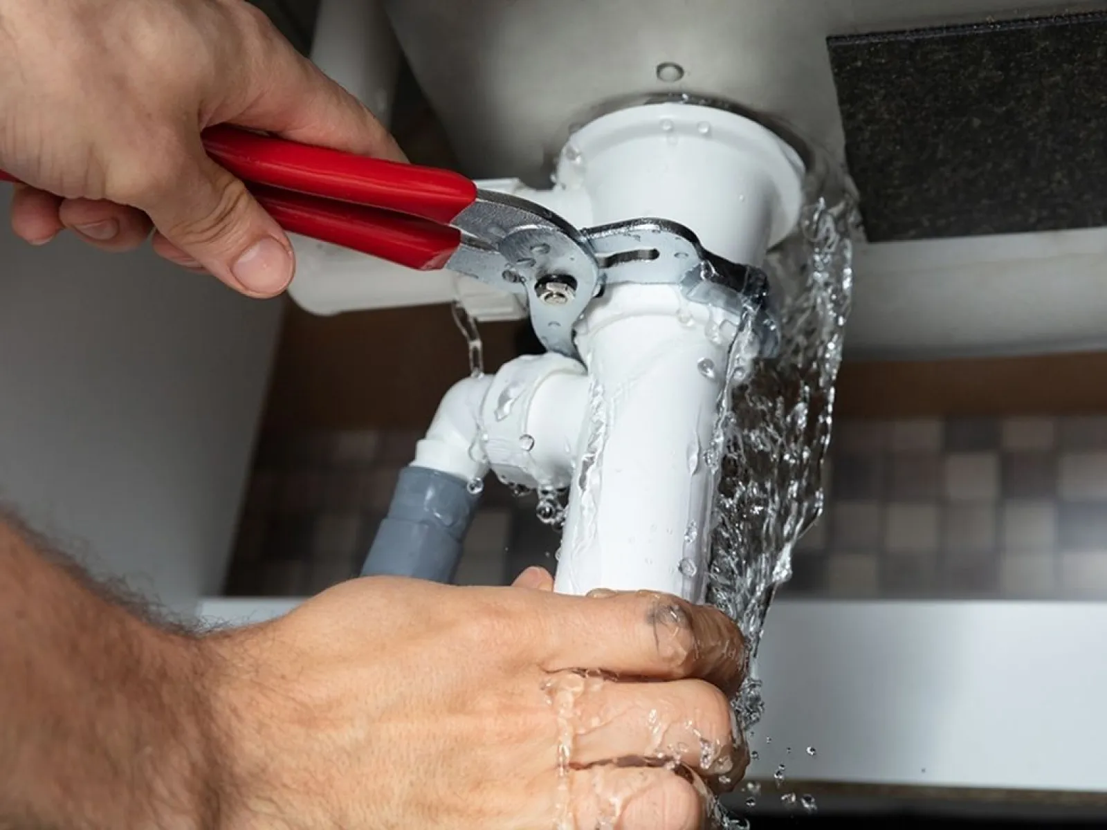 Hands using pliers to fix leaking white pipe under sink with water spraying out.